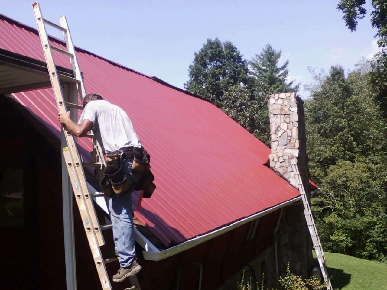 Roofer on a ladder beside a red metal roof and stone chimney — Soffit & Fascia Repair by Roofing Brothers in Camp Murray